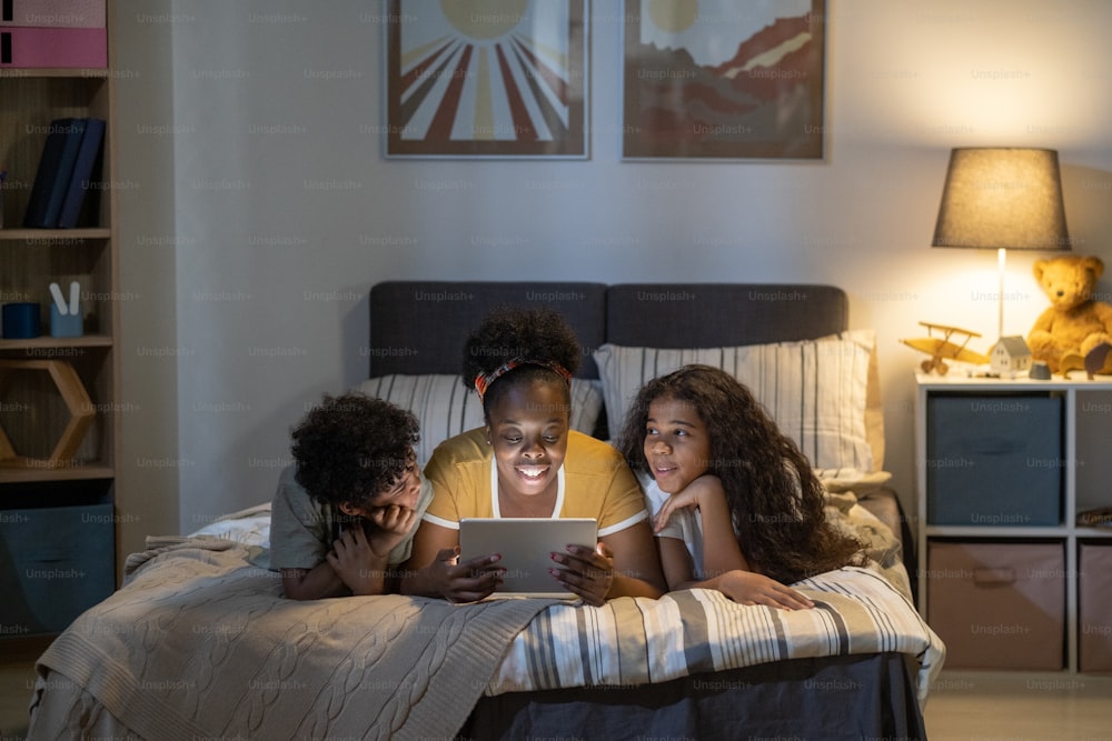 Happy curly-haired African American mother lying on bed and using tablet while reading fairy tale to kids before bedtime