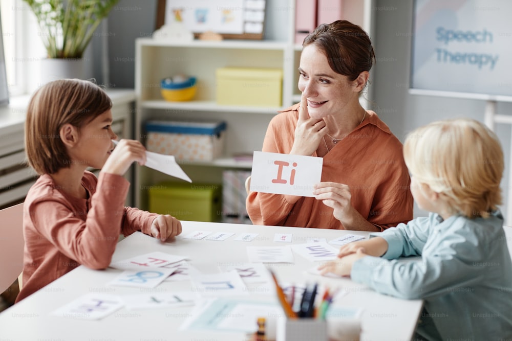 Young woman sitting at the table with children showing the card with letter and teaching them to read