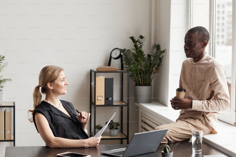 Side view portrait of two smiling business people communicating at break in minimal office interior