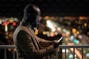 Side view of African businessman with smartphone texting on balcony of office building at night