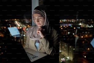 Jeune femme d’affaires sérieuse en hijab réseautage par fenêtre dans un grand bureau dans la nuit