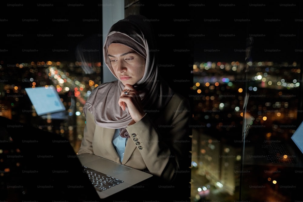 히잡을 쓴 젊은 진지한 사업가 밤에 큰 사무실에서 창문으로 네트워킹을 하고 있다