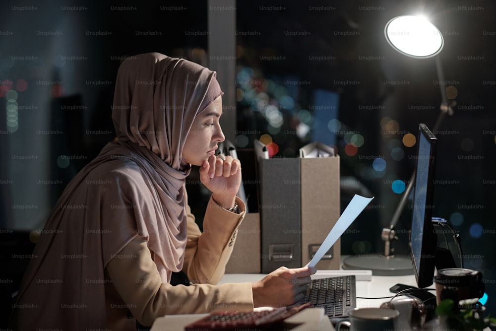 Side view of young businesswoman in hijab looking through papers by workplace in office at night time