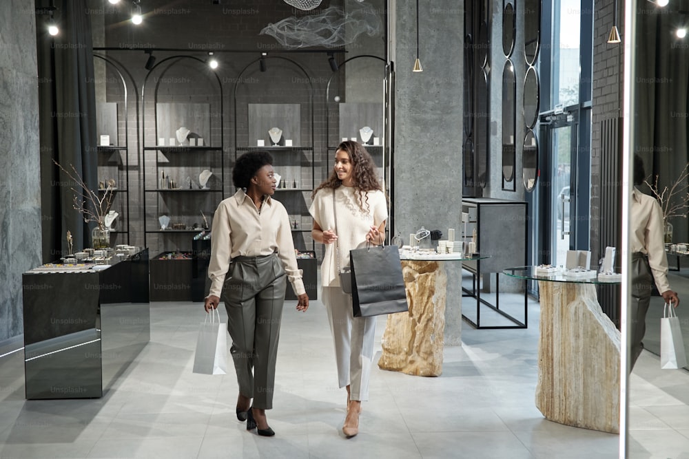 Mujeres jóvenes interculturales con ropa casual elegante que se mueven a lo largo de la joyería o boutique mientras compran en un centro comercial en Black Friday