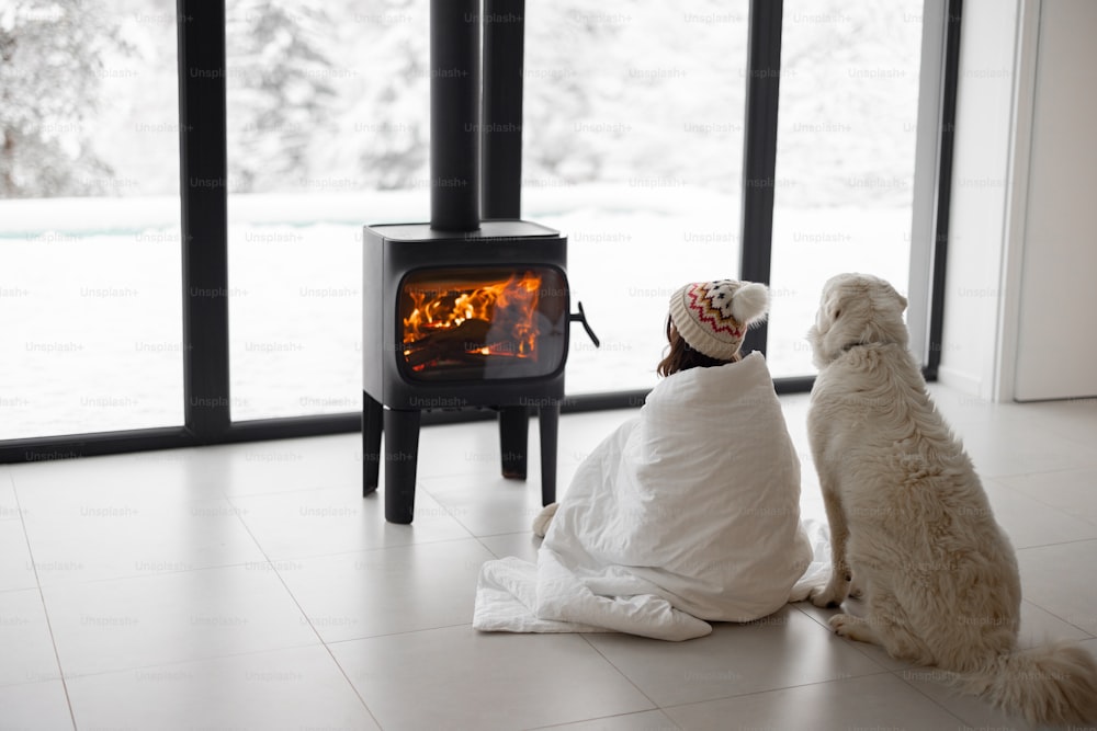 Woman sitting with her dog by the fireplace at modern house on nature during winter time. Concept of winter mood and comfort at home. Girl in hat covered with blanlet