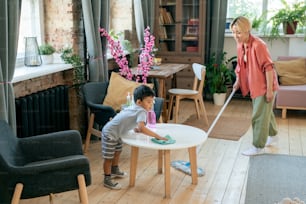 Junge asiatische Frau, die den Boden reinigt, während ihr entzückender kleiner Sohn den Tisch mit Desinfektionsmittel im Wohnzimmer abwischt
