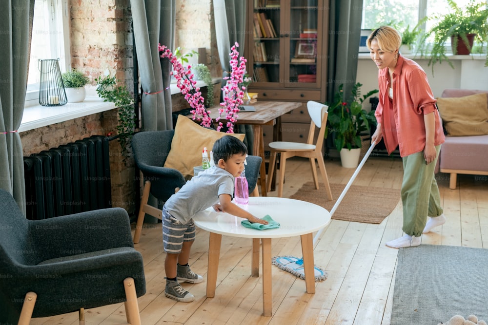Joven asiática limpiando piso mientras su adorable hijo pequeño limpia la mesa con desinfectante en la sala de estar