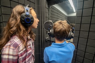 Ein junges Mädchen und ein Junge mit Kopfhörern stehen vor dem Mikrofon in einem schalldichten Raum