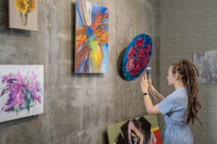 Young woman standing in front of the wall with modern art and using her mobile phone to make photos