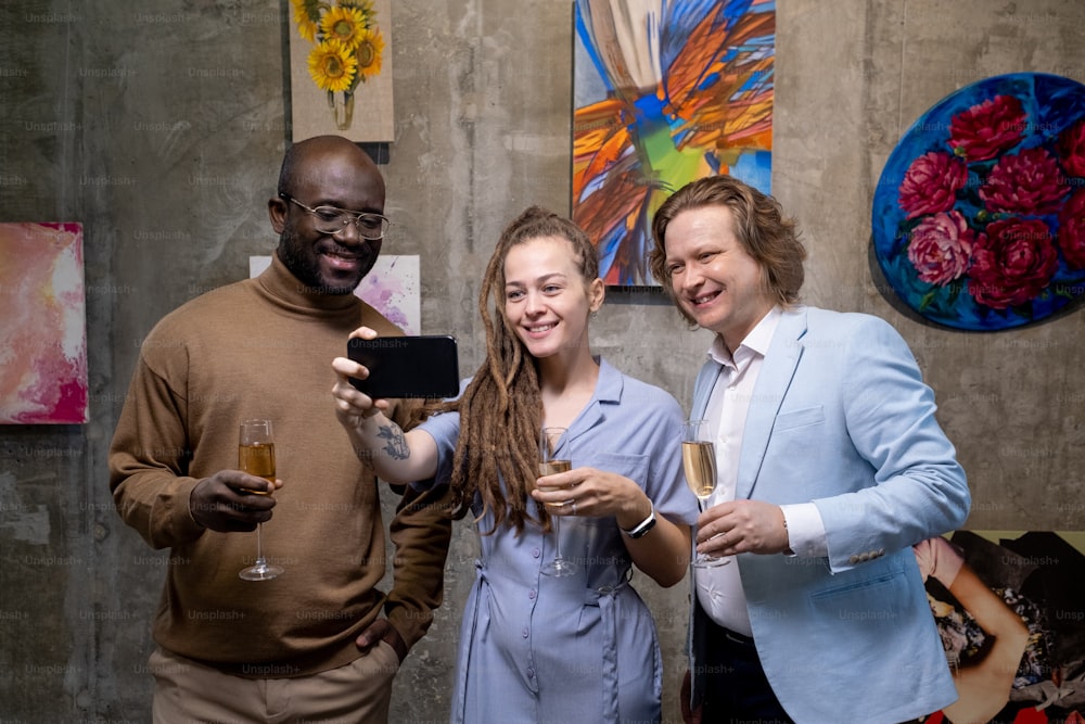 Group of happy people with champagne making selfie portrait on mobile phone against the paintings while visiting exhibition in art gallery