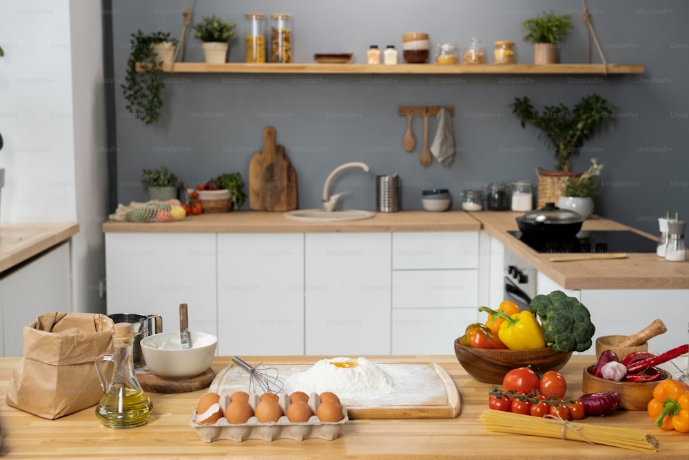 Interior of kitchen with wooden table, fresh vegetables, sifted flour, eggs, bottle of olive-oil and bunch of spaghetti