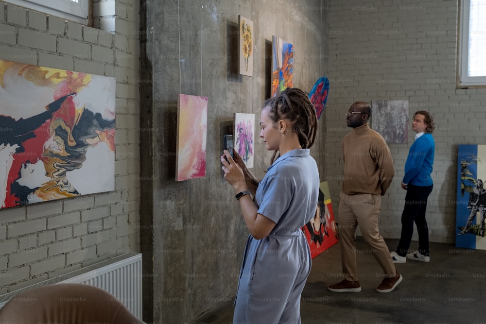 Jeune femme photographiant les peintures sur son téléphone portable lors de sa visite dans une galerie d’art avec d’autres personnes en arrière-plan