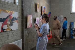Giovane donna che fotografa i dipinti sul suo telefono cellulare durante la sua visita in galleria d'arte con altre persone sullo sfondo