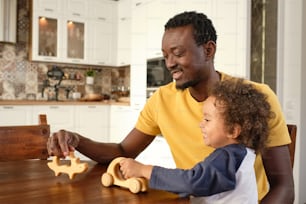 Pai feliz e filho pequeno bonito jogando carrinhos de brinquedo de madeira e desfrutando de lazer enquanto sentado à mesa na cozinha