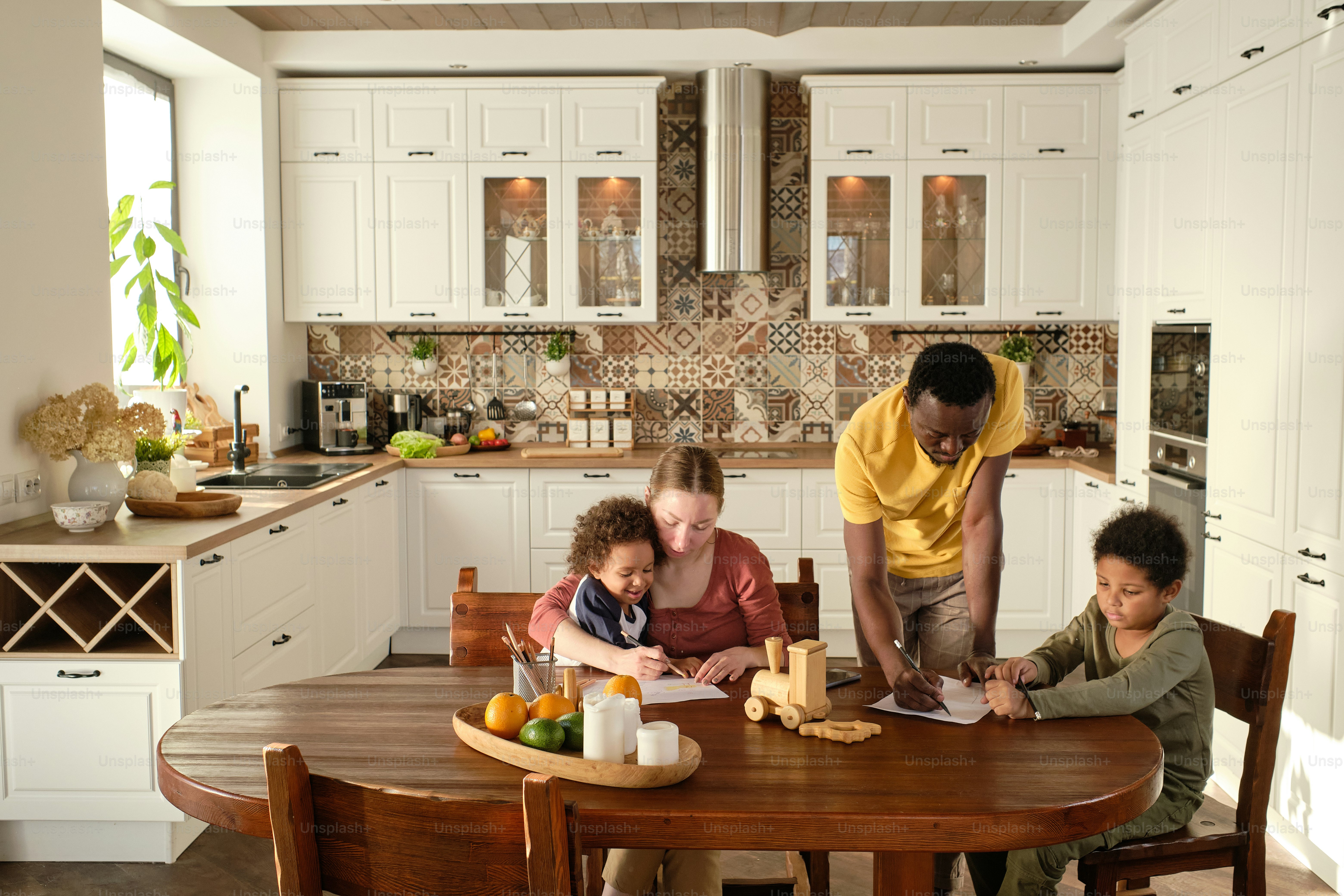 Dos jóvenes padres interculturales enseñan a sus lindos hijos pequeños a dibujar mientras están reunidos junto a una mesa de madera en la cocina