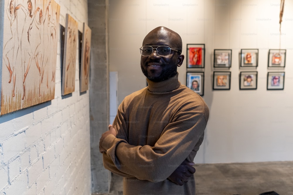 Ritratto di giovane visitatore africano che sorride alla macchina fotografica mentre è in piedi con le braccia incrociate vicino al muro con opere d'arte nel museo d'arte