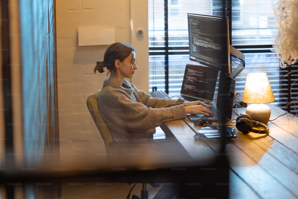 Une jeune femme travaille sur des ordinateurs, assise sur son lieu de travail à l’intérieur confortable du bureau à domicile. Concept de freelance et de travail à distance depuis à domicile. Programmeur écrivant du code. Femme de race blanche portant des vêtements domestiques.
