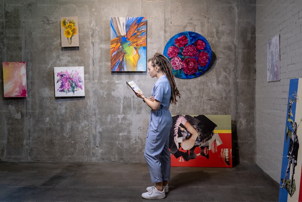 Mulher nova lendo um livreto enquanto está na galeria de arte com belas pinturas na parede