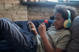 롤리팝과 스마트폰을 들고 거실에서 여유롭게 소파에 누워 온라인 영화를 보는 행복한 귀여운 아프리카 소년