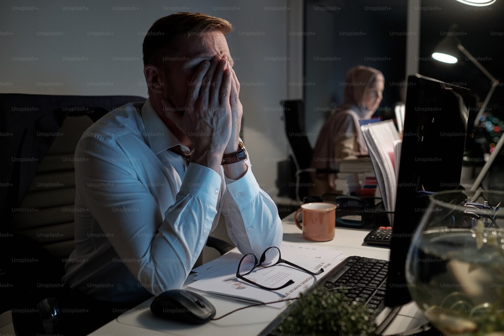 Exhausted businessman in white shirt keeping his arms on face while working in front of computer in office late at night