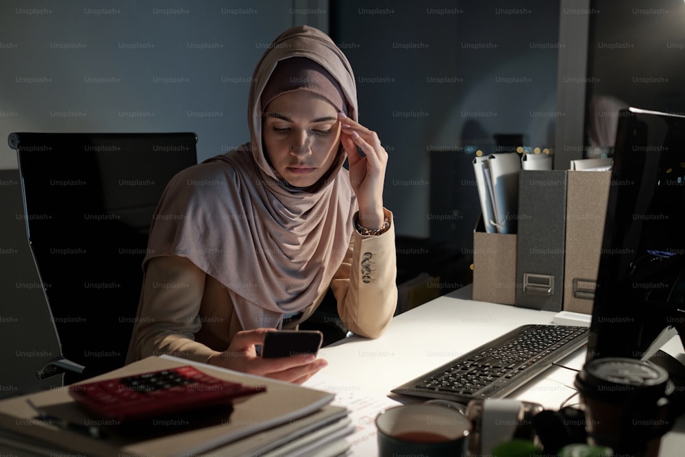 히잡을 쓴 젊은 진지한 사업가가 컴퓨터 앞 책상 옆에 앉아 있는 동안 밤늦게 스마트폰으로 스크롤한다