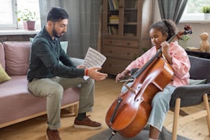 Menina bonita tocando violoncelo enquanto seu professor de música sentado por perto e explicando como tocar instrumento musical