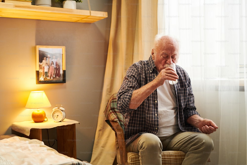 寝室の肘掛け椅子に座って水を飲み、ガラスから、朝に薬を飲む老人