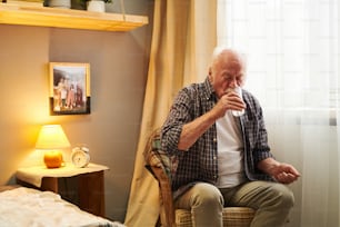 Älterer Mann sitzt auf einem Sessel in seinem Schlafzimmer und trinkt Wasser aus dem Glas, er nimmt morgens Tabletten