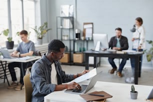 African young businessman sitting at the table reading a document and typing it on laptop during his work at office