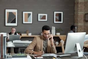 Young contemporary businessman drawing picture of interior and talking to client on mobile phone while sitting by workplace