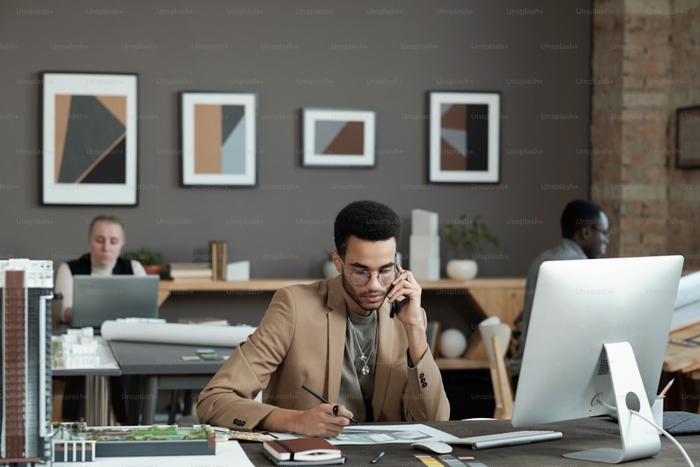 Junger zeitgenössischer Geschäftsmann, der ein Bild des Interieurs zeichnet und mit dem Kunden auf dem Handy spricht, während er am Arbeitsplatz sitzt