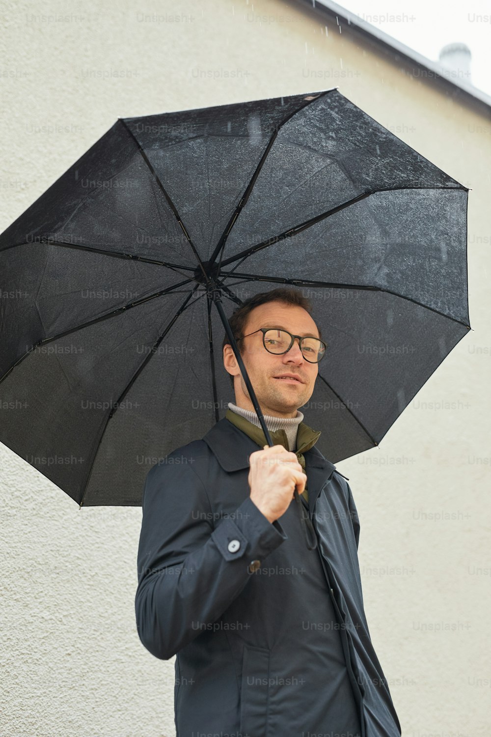 Vertical low angle medium shot of handsome Caucasian man wearing eyeglasses standing outdoors under umbrella looking at camera