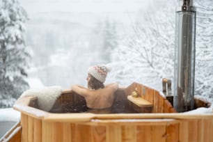 Femme se relaxant dans un bain chaud à l’extérieur, assise et profitant d’une vue magnifique sur les montagnes enneigées. Vacances d’hiver à la montagne, concept de soins à l’eau chaude. Femme de race blanche portant un chapeau d’hiver