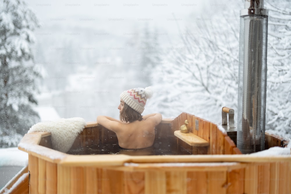 Donna che si rilassa in bagno caldo all'aperto, seduta e godendo di una splendida vista sulle montagne innevate. Vacanze invernali in montagna, concetto di trattamento dell'acqua calda. Donna caucasica che porta il cappello invernale