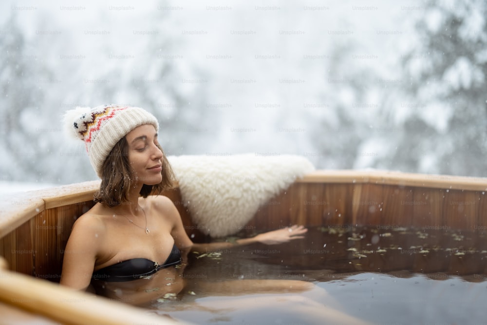 Donna che si rilassa nel bagno caldo all'aperto, godendo di un centro termale sulle montagne innevate. Vacanze invernali in montagna, concetto di trattamento dell'acqua calda. Donna caucasica che porta il cappello invernale