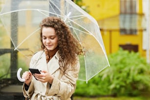 Ritratto medio orizzontale della giovane donna che indossa il trench in piedi all'aperto sotto l'ombrello sul giorno di pioggia che naviga in Internet sullo smartphone