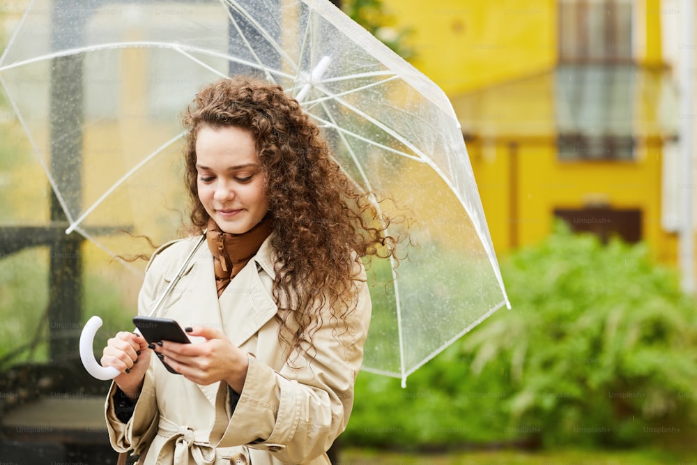 Retrato horizontal mediano de una mujer joven con gabardina parada al aire libre bajo paraguas en un día lluvioso navegando por Internet en un teléfono inteligente