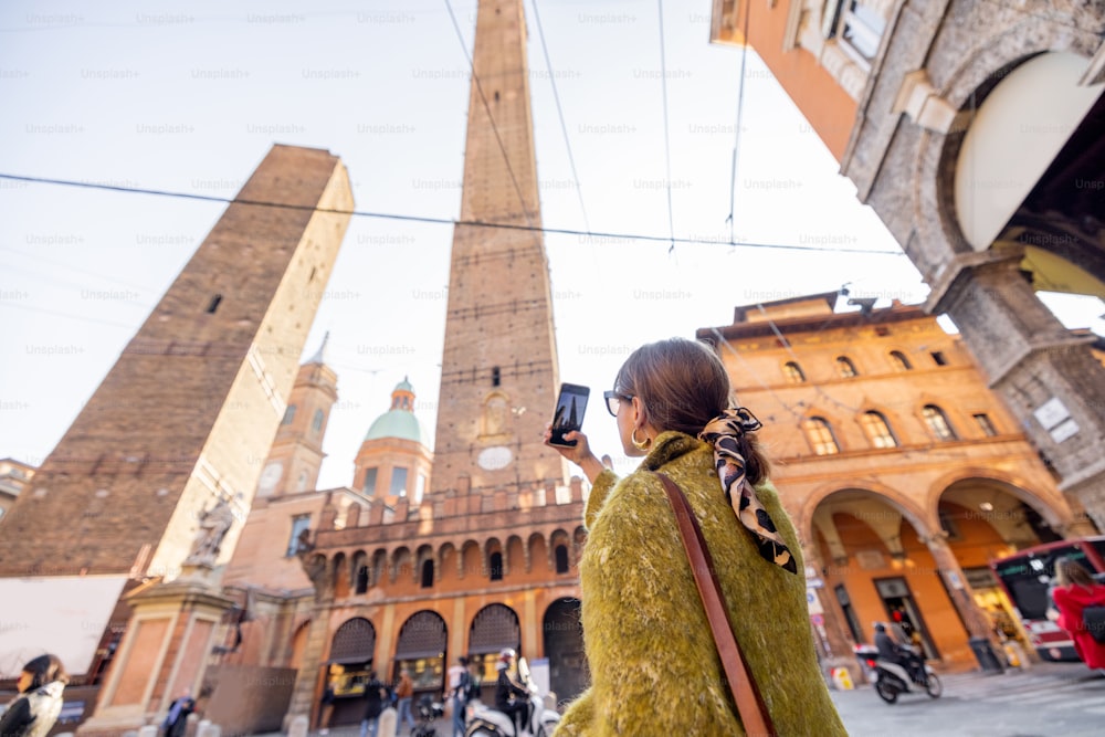 Woman photographing on phone famous Asinelli towers while traveling in Bologna city. Concept of visiting Italian landmarks in Emilia Romagna region