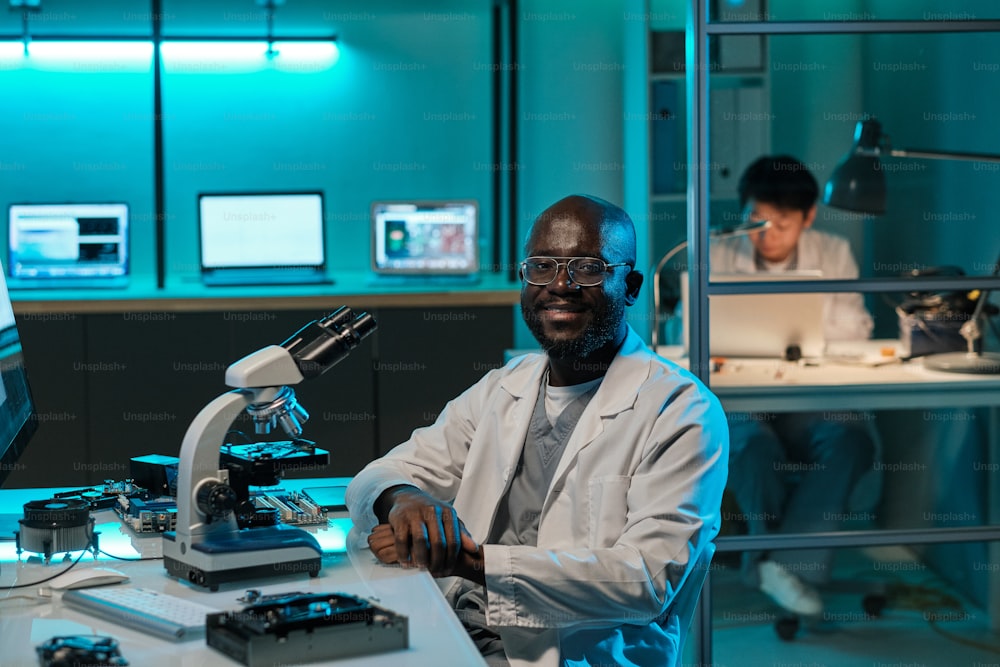 Giovane scienziato africano felice con il microscopio seduto sul posto di lavoro contro la collega femminile e guardando la macchina fotografica