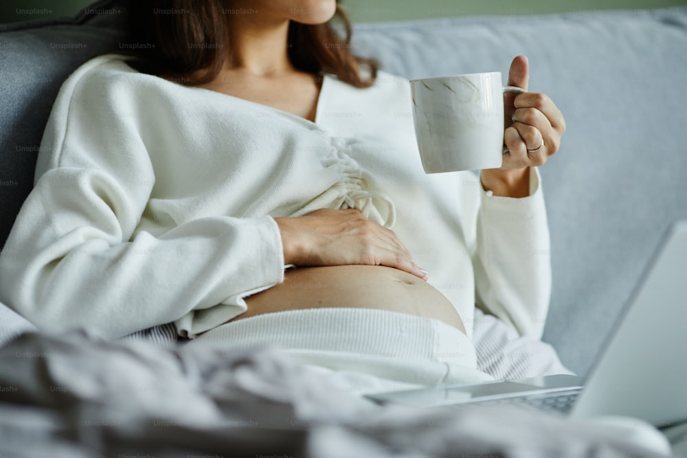 Close-up of young pregnant woman in pyjamas drinking tea while resting in her bed