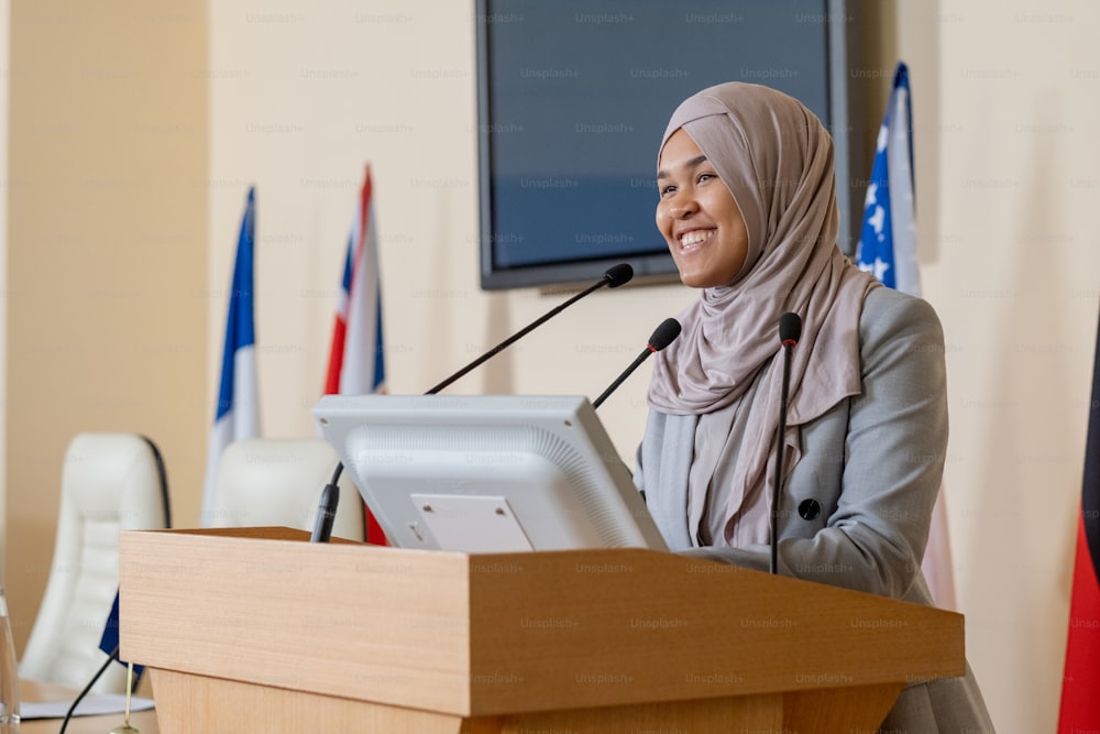 히잡을 쓴 젊은 성공한 무슬림 사업가가 회의에서 외국 동료들을 위해 연설하는 동안 트리뷴 옆에 서 있다