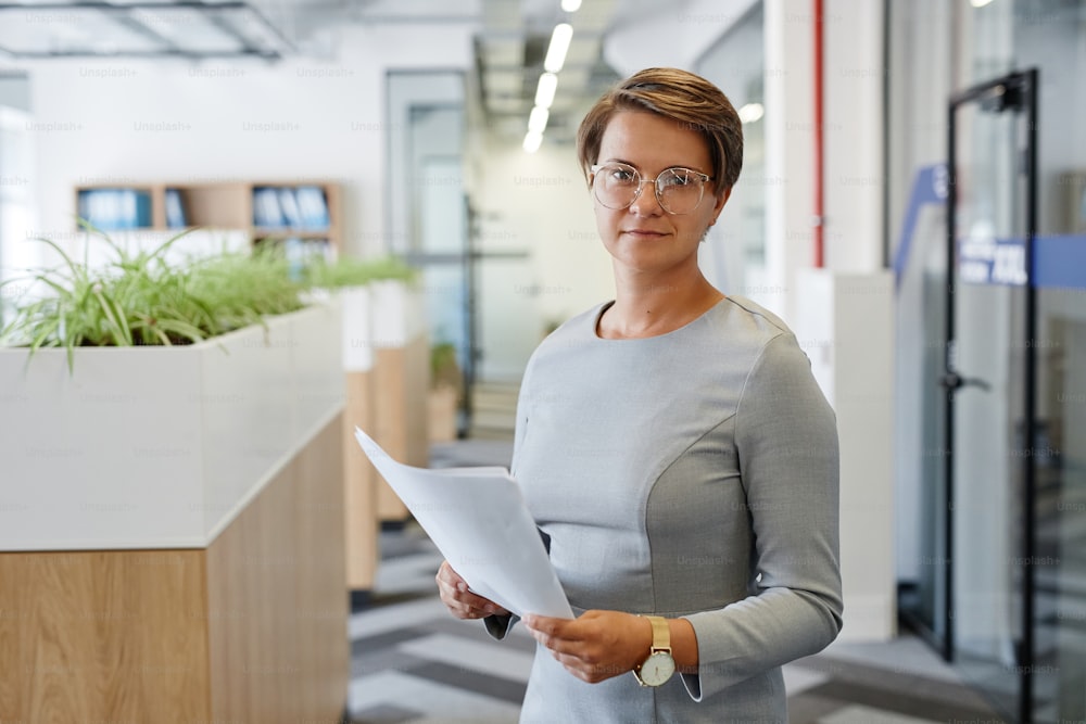 Retrato de cintura para arriba de gerente femenino adulto mirando a la cámara mientras está de pie en la oficina y sosteniendo el documento, espacio de copia
