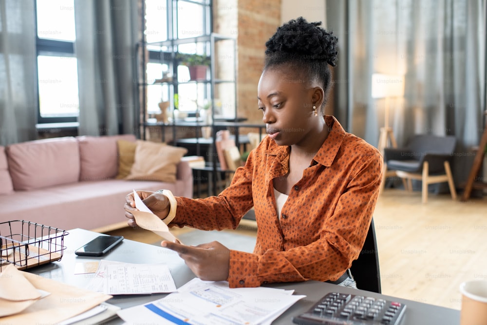 Young African woman sitting by table and putting filled form into envelope before sending it to organization