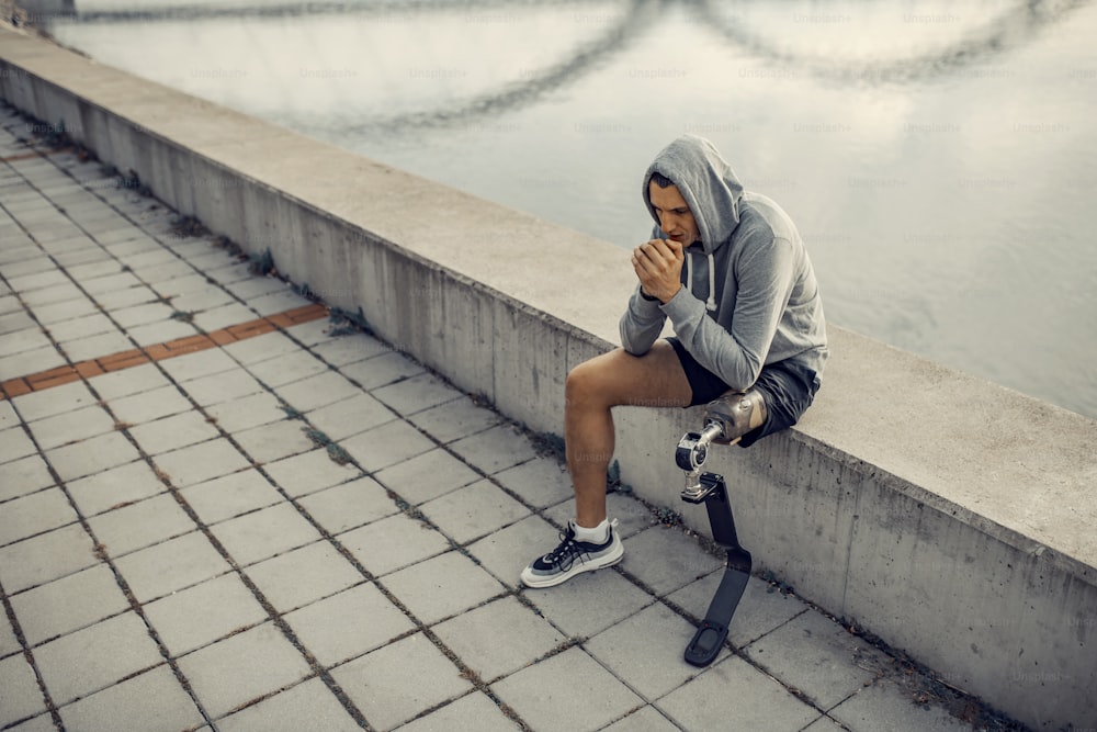 A handicapped sportsman with artificial leg with hooded sweatshirt sitting at the quay and warming up hands.