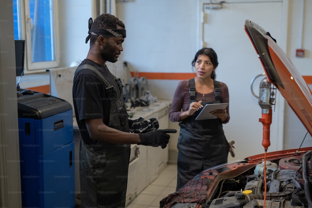 Retrato de vista lateral de dos mecánicos reparando automóviles en un taller de automóviles con enfoque en el trabajador afroamericano inspeccionando el motor, espacio de copia