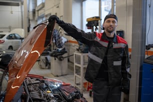 Mecánico joven en ropa de trabajo que abre la cubierta del capó del automóvil mientras está parado junto al vehículo en el garaje o taller