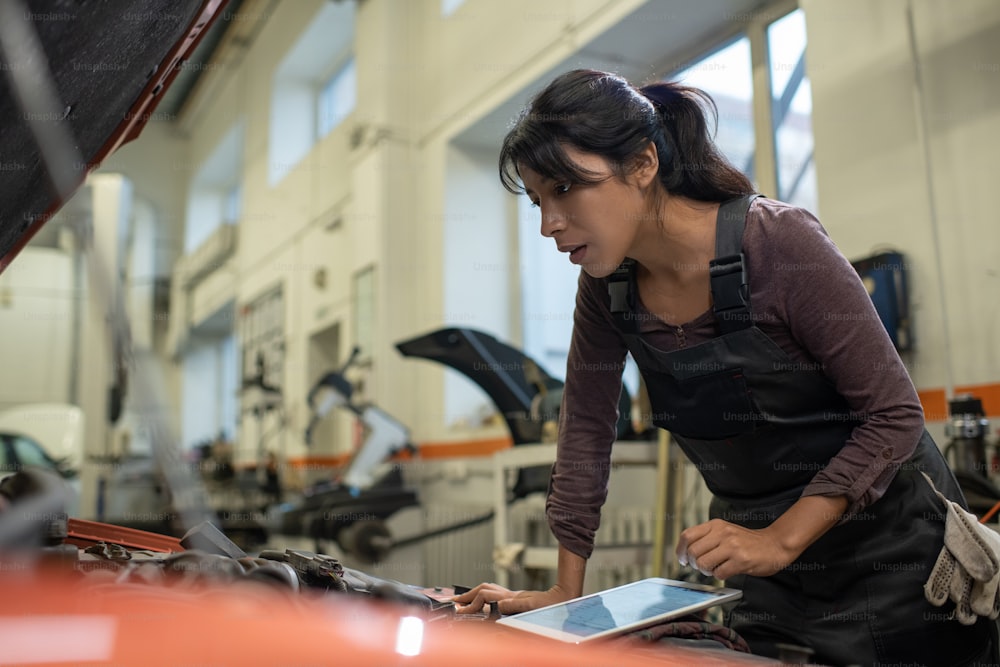 自動車修理工場で車両を検査しながらボンネットの下を見る若い女性整備士の側面図のポートレート、コピー用スペース