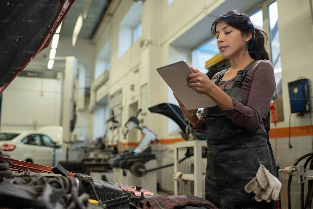 Young Hispanic woman in overalls looking at screen of digital tablet in her hands while repairing broken motor of car