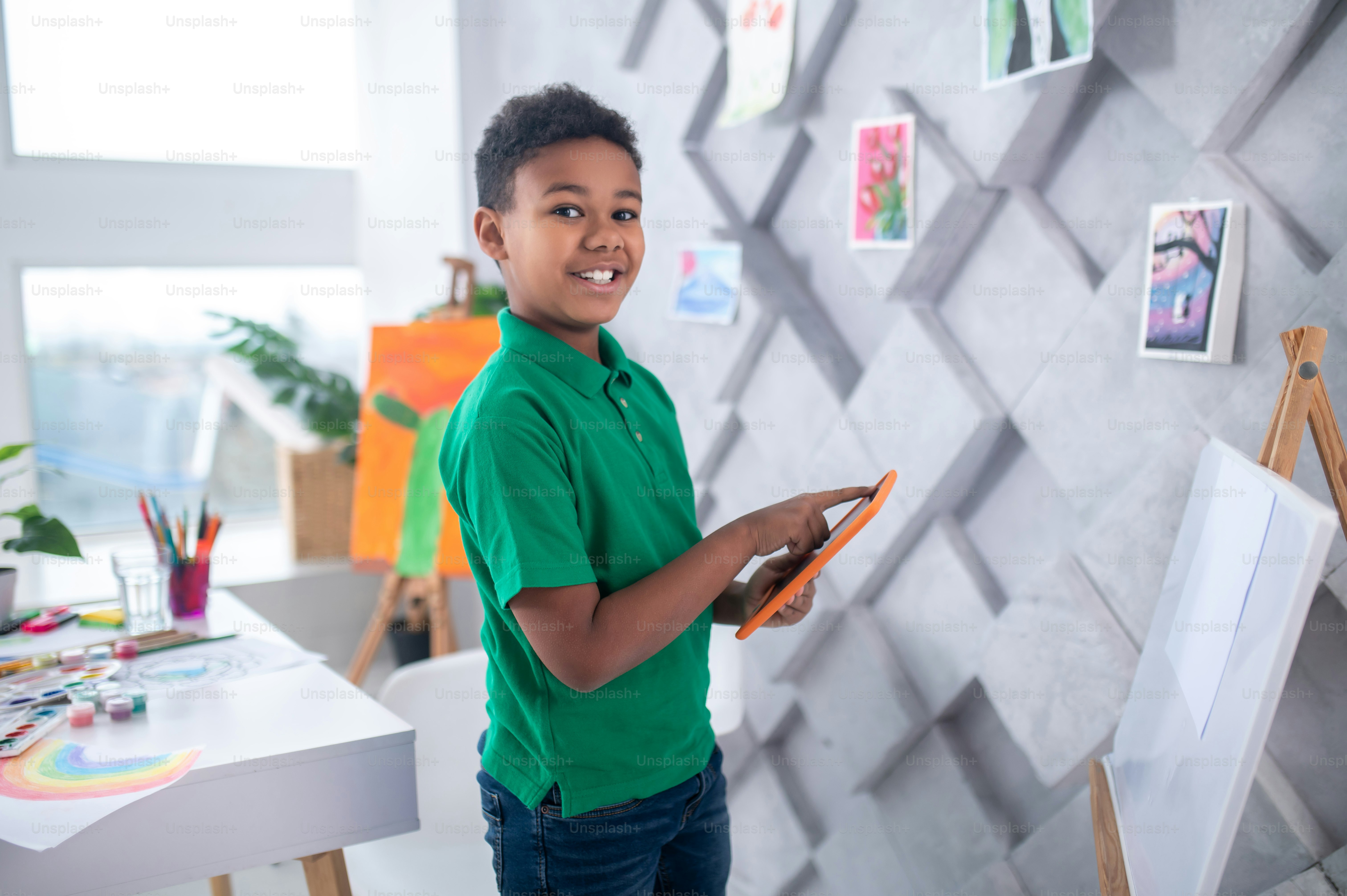 Idea. Alegre niño de piel oscura en edad escolar con camiseta verde tocando la pantalla de la tableta con el dedo parado cerca del caballete de dibujo mirando a la cámara en la habitación de luz