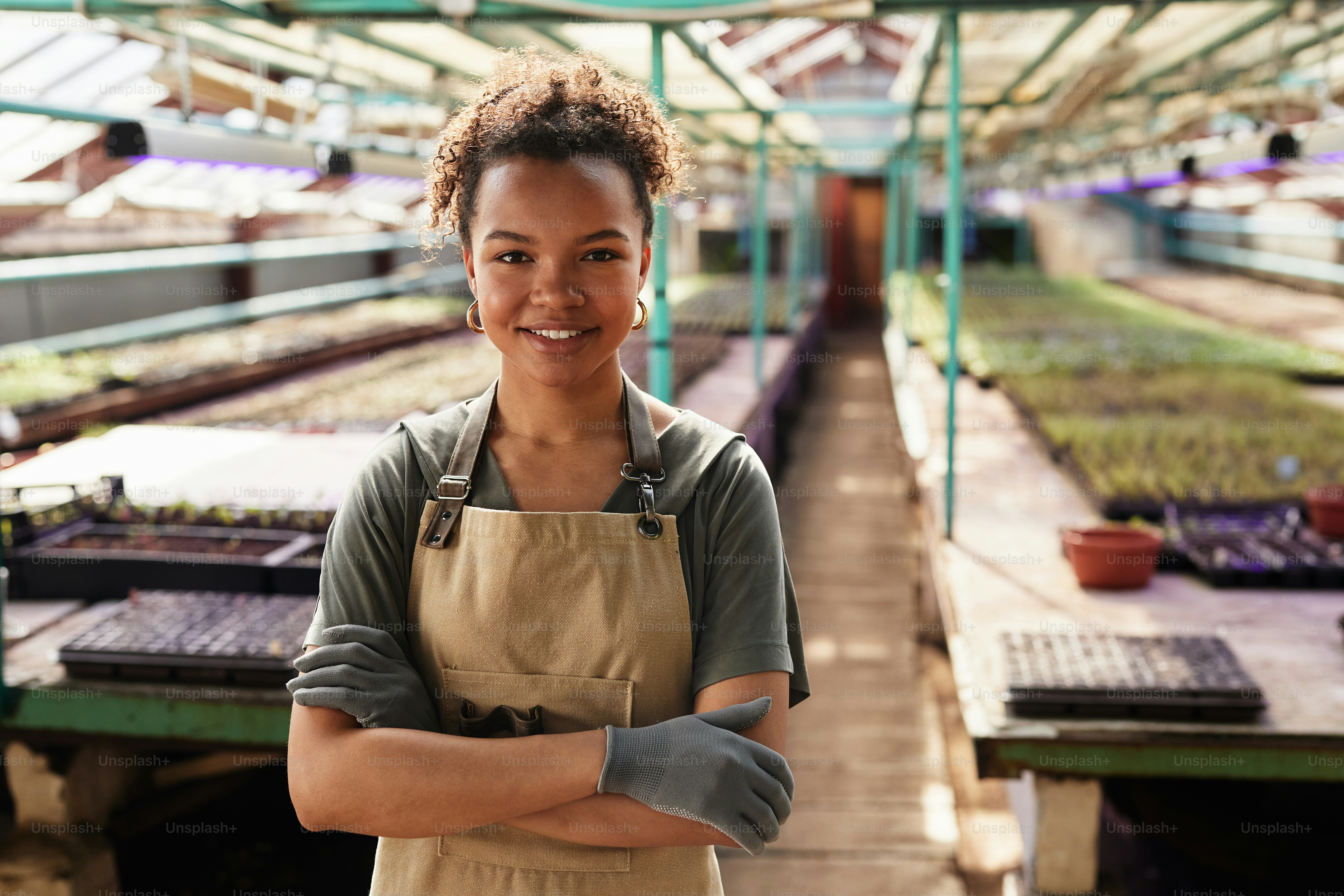 Feliz joven trabajadora afroamericana de jardinería en ropa de trabajo y guantes protectores cruzando sus brazos por el pecho